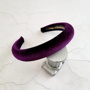 Può includere: Un'ampia fascia per capelli in velluto viola. La fascia è su una superficie bianca con una base argentata.