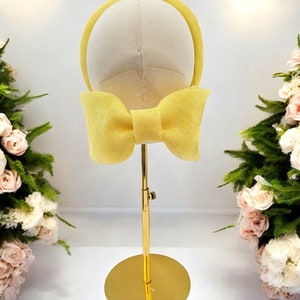 May include: A yellow headband with a large bow on a gold stand. The headband is made of a woven material.