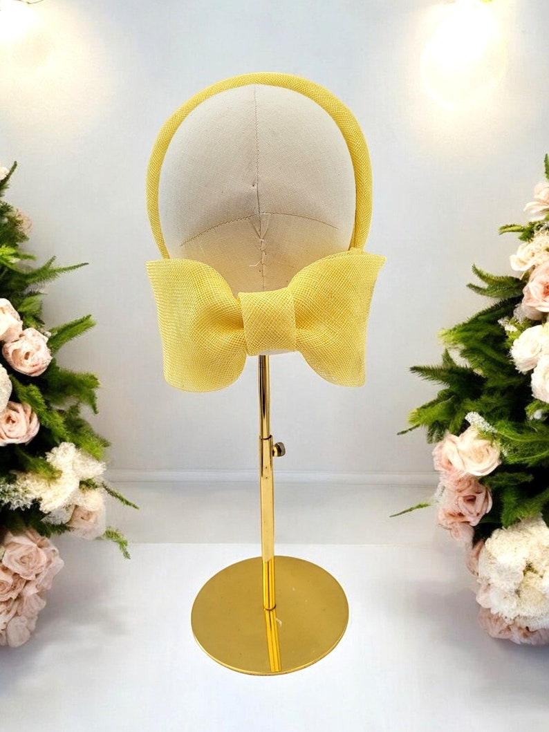 May include: A yellow headband with a large bow on a gold stand. The headband is made of a woven material.