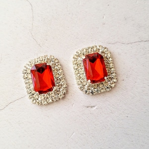 以下が含まれることがあります： 長方形の赤い宝石装飾が2つ、銀色のラインストーンの縁取り付き。宝石は鮮やかな赤色で、ラインストーンが輝いています。さまざまなクラフトプロジェクトに適しています。