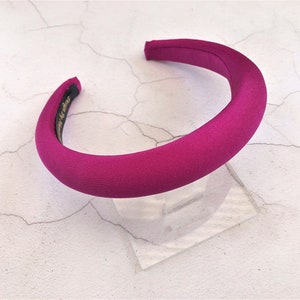 Può includere: Un'ampia fascia per capelli di colore rosa fucsia realizzata in tessuto. La fascia è su una superficie bianca.