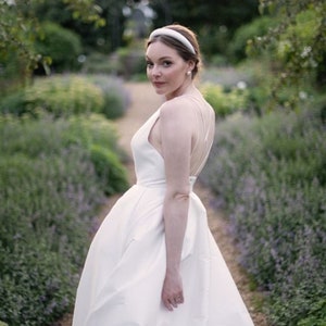 May include: A bride wearing a white wedding dress with a simple, elegant design. She is standing in a garden with purple flowers and greenery.