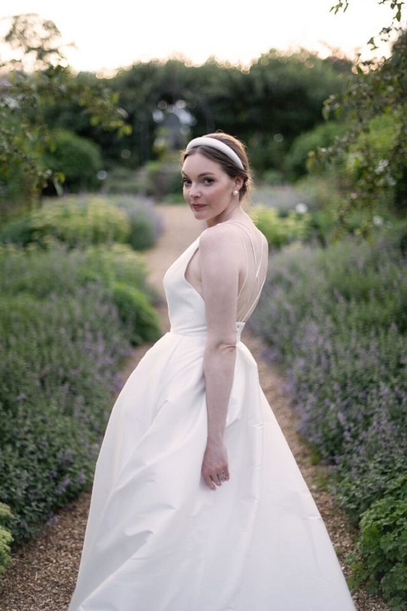 May include: A bride wearing a white wedding dress with a simple, elegant design. She is standing in a garden with purple flowers and greenery.