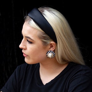 May include: A woman with blonde hair wearing a black headband and black and white patterned earrings. She is wearing a black t-shirt.