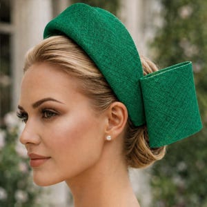 Può includere: Una fascia verde brillante in tessuto strutturato, con un dettaglio rettangolare sul lato. La fascia è indossata da una persona con capelli biondi e un orecchino di perla. Lo sfondo è sfocato, suggerendo un ambiente esterno.