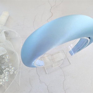 Può includere: Un cerchietto in raso blu chiaro con una fascia larga e imbottita. Il cerchietto è esposto su un supporto in plastica trasparente.