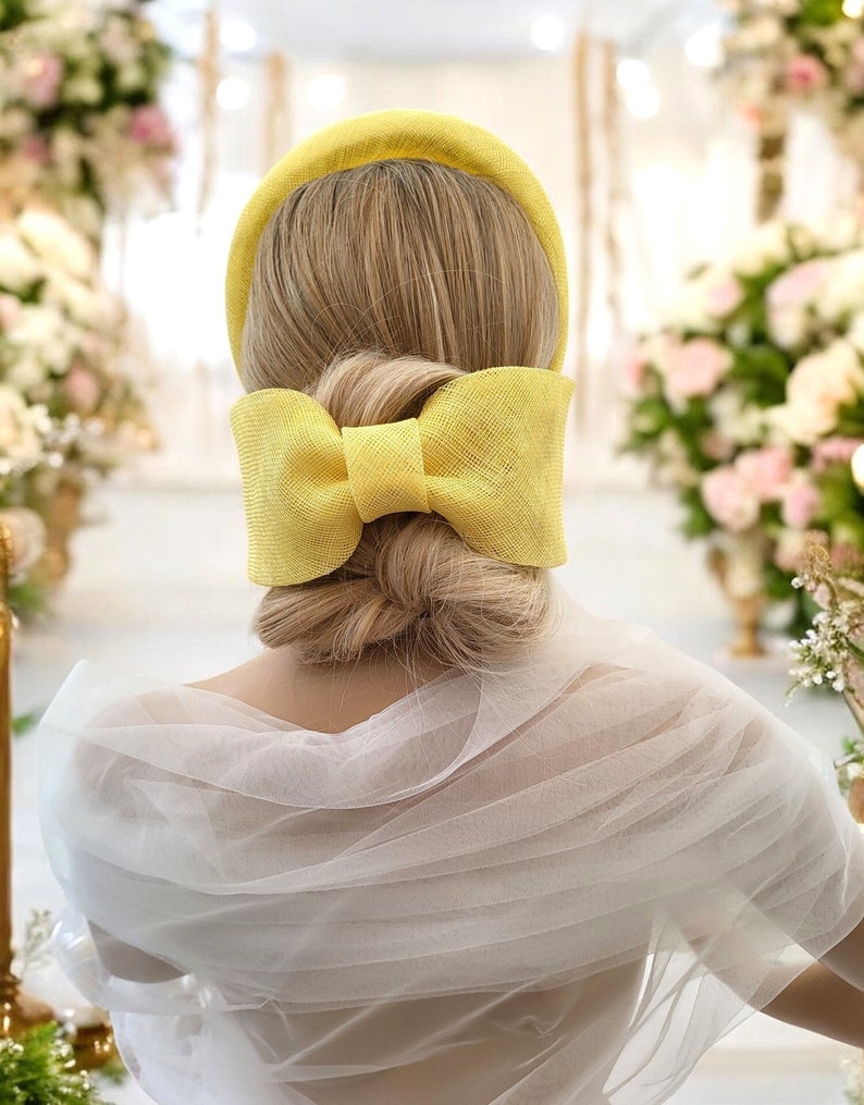 May include: A yellow headband with a large bow at the back. The headband is made of a woven material and is being worn by someone with blonde hair.