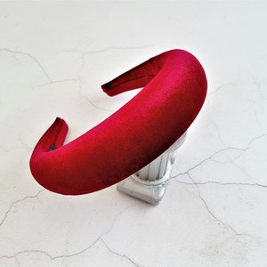 Può includere: Una fascia imbottita in velluto rosso intenso. La fascia è spessa e curva, progettata per adattarsi comodamente alla testa. L'accessorio è esposto su una superficie bianca con un motivo simile al marmo.