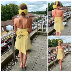 May include: A woman wearing a yellow strapless dress with a large yellow bow at the back. She is also wearing a yellow headband with a bow on top. The dress is short and has a fitted bodice. The woman is standing on a balcony with a railing.