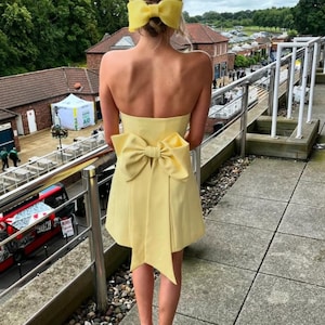 May include: A yellow strapless dress with a large bow at the back. The dress is short and has a fitted bodice. The model is wearing a yellow headband with a bow on top.