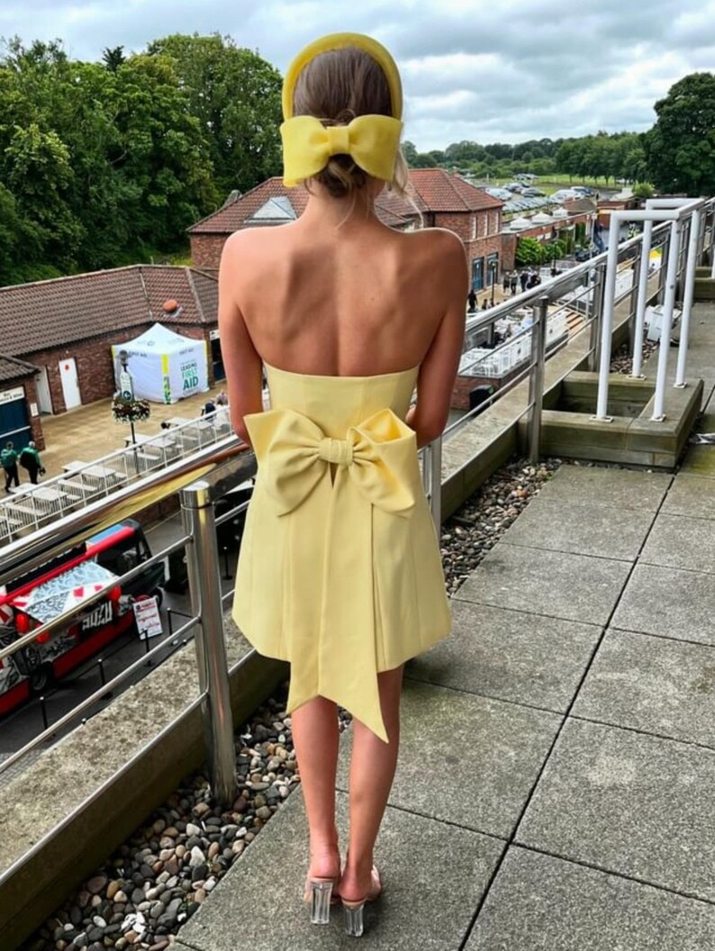 May include: A yellow strapless dress with a large bow at the back. The dress is short and has a fitted bodice. The model is wearing a yellow headband with a bow on top.