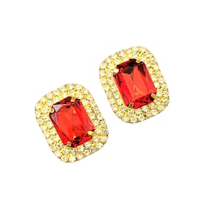 Puede incluir: Un par de pendientes de tono dorado con piedras preciosas rectangulares rojas rodeadas de diamantes de imitación transparentes.