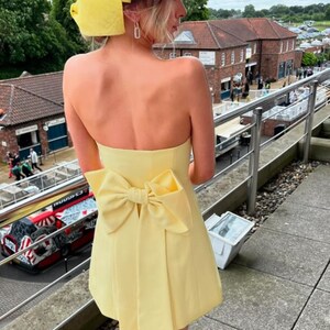 May include: A yellow strapless dress with a large bow at the back. The dress is short and has a fitted bodice. The model is wearing a yellow headband.