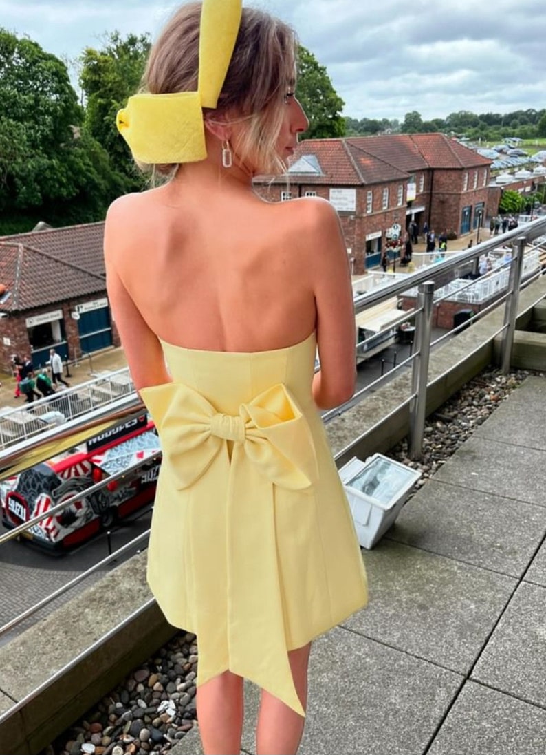 May include: A yellow strapless dress with a large bow at the back. The dress is short and has a fitted bodice. The model is wearing a yellow headband.