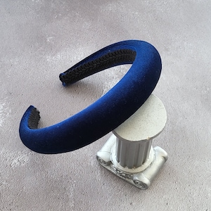 Può includere: Un'ampia fascia per capelli in velluto blu con un bordo intrecciato nero. La fascia è appoggiata su un piccolo piedistallo decorativo argentato.