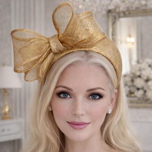 May include: A gold-coloured headband featuring a large bow made of a sheer, textured fabric. The headband is worn by a person with blonde hair and blue eyes. The background includes a mirror and a white wall.