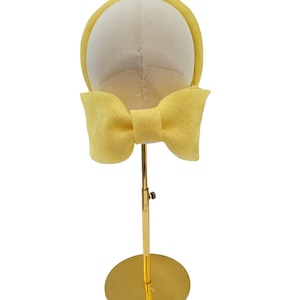 May include: A yellow headband with a large bow at the back. The headband is displayed on a gold stand.