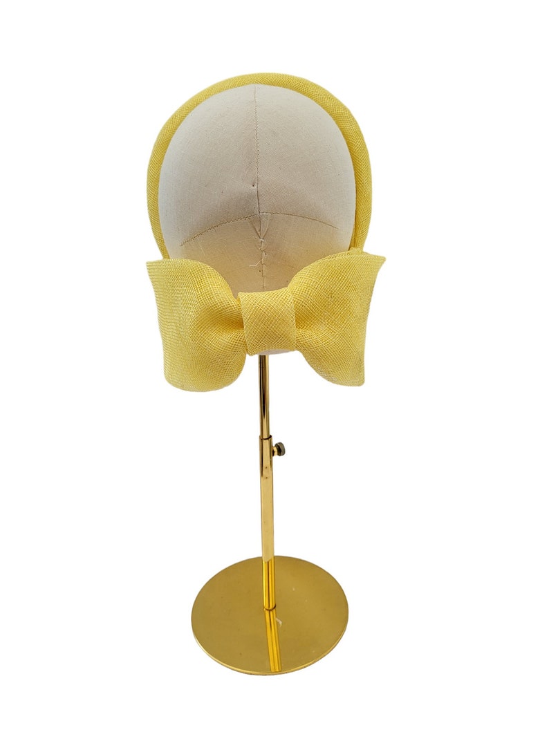 May include: A yellow headband with a large bow at the back. The headband is displayed on a gold stand.