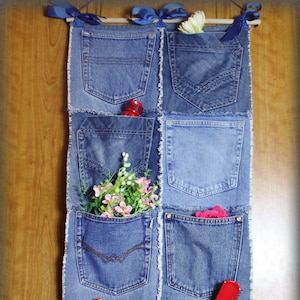 Puede incluir: Un organizador de pared de mezclilla con ocho bolsillos, cada uno hecho de un par diferente de jeans. Los bolsillos están decorados con flores, un juguete rojo, un peine rojo y un juguete blanco.