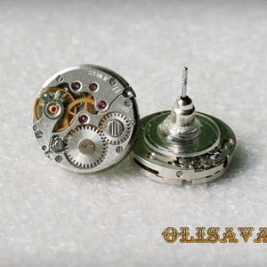 Puede incluir: Un par de pendientes de plata con un diseño steampunk. Cada pendiente presenta una esfera de reloj circular con engranajes expuestos y una pequeña joya rosa. Los pendientes están unidos a un poste de plata.