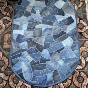 Op de afbeelding: Ovaal tapijt gemaakt van denim lappen in verschillende blauwtinten. Het patchwork ontwerp heeft verschillende denim wassingen, wat een textuureffect creëert. Het tapijt heeft een blauwe rand. Afmetingen: ongeveer 150 cm x 90 cm.