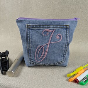 Op de afbeelding: Een blauwe denim ritszak met een paarse rits en een roze geborduurde letter "J" aan de voorkant. De tas ligt op een beige oppervlak met een zwarte haarspeld en een zilveren buis.