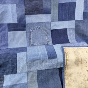 Op de afbeelding: Een patchworkquilt gemaakt van verschillende tinten blauwe denim. De quilt heeft een verscheidenheid aan rechthoekige en vierkante patches, waaronder een denim zak. De verschillende tinten blauw creëren een textuurrijk en visueel interessant ontwerp.
