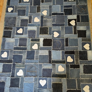 Peut inclure: Une courtepointe en patchwork faite de tissu en jean avec des appliques en forme de cœur blanc. La courtepointe est composée de différentes nuances de jean bleu et a un bord effiloché.