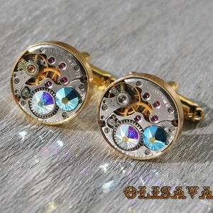 May include: A pair of gold cufflinks with a round, silver, watch-like design. The cufflinks feature intricate gears and two clear, iridescent crystals.
