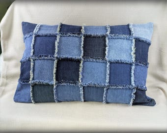 May include: Rectangular denim pillow with a patchwork design. The pillow features squares of various shades of blue denim, creating a textured and visually appealing pattern. The edges of the squares have a frayed, light-colored trim.