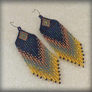 May include: A pair of beaded earrings with a diamond-shaped top and long fringe. The earrings feature a gradient of colours, including navy blue, peach, light blue, and gold. The earrings are made with small, iridescent beads.