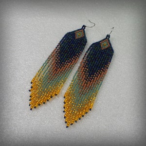 May include: A pair of long, beaded earrings with a gradient of colors, starting with dark blue at the top and transitioning to gold at the bottom. The earrings are made with small, round beads and have a geometric design at the top.