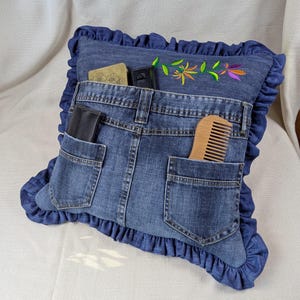 May include: A square denim cushion featuring a ruffled border and two functional pockets made from jeans. The cushion is decorated with an embroidered floral design in yellow, green, and purple. A wooden comb and a black wallet are tucked into the pockets.