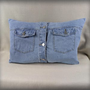 Puede incluir: Un cojín rectangular decorativo que simula una chaqueta vaquera azul claro. Tiene dos bolsillos delanteros y se abrocha con botones plateados. El cojín está sobre una superficie de color neutro.