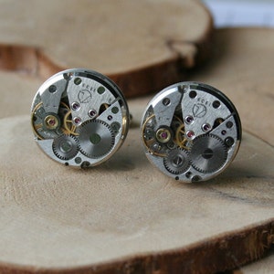 Peut inclure: Une paire de boutons de manchette argent&eacute;s avec un design circulaire mettant en valeur des engrenages de montre apparents et un petit bijou rose. Les boutons de manchette sont sur une surface en bois.