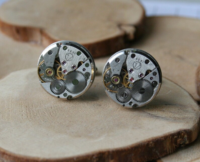 Peut inclure: Une paire de boutons de manchette argent&eacute;s avec un design circulaire mettant en valeur des engrenages de montre apparents et un petit bijou rose. Les boutons de manchette sont sur une surface en bois.