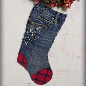 May include: A denim Christmas stocking with a red plaid heel and toe. The blue jean material has decorative embroidery near the pocket. The stocking is hanging on a white wall with a Christmas garland above it.