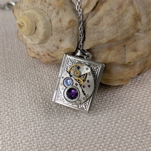 Op de afbeelding: Een zilverkleurige rechthoekige hangerketting met ingewikkelde details. De hanger heeft een miniatuur uurwerkontwerp met tandwielen, een paarse edelsteen en blauwe en rode accenten. De ketting hangt aan een fijne zilveren ketting.