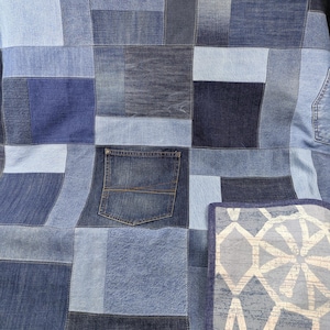 Op de afbeelding: Een patchworkquilt gemaakt van verschillende tinten blauwe denim. De quilt heeft een verscheidenheid aan denim vierkanten en rechthoeken, waaronder een denim zak. Een kussen met patroon met een blauwe rand is ook zichtbaar.
