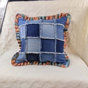 Puede incluir: Un cojín cuadrado con un diseño de retazos de varios cuadrados de tela de mezclilla azul con bordes deshilachados. El cojín está bordeado por un volante con un patrón floral multicolor en tonos naranja, verde y azul.