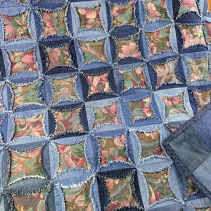 Könnte beinhalten: Eine handgefertigte Steppdecke mit einem sich wiederholenden Muster aus blauen Jeansstoffquadraten mit ausgefransten Kanten, die sich mit quadratischen Flicken aus Blumenstoff in Rosa-, Grün- und Brauntönen abwechseln. Die Jeansstoffteile bilden ein strukturiertes Gitter.
