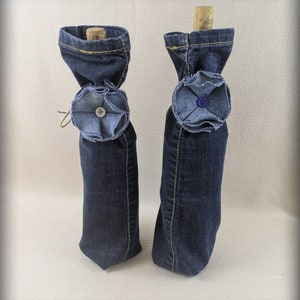 May include: Two denim wine bottle covers. Each cover is made from dark blue denim and features a decorative denim flower with a button center. The tops of the covers are folded over, and a cork is visible in each.