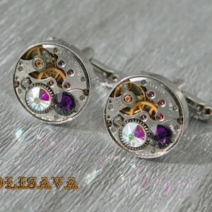 Könnte beinhalten: Ein Paar silberne Manschettenknöpfe mit Steampunk-Design. Jeder Manschettenknopf verfügt über ein rundes Zifferblatt mit komplizierten Zahnrädern, einem violetten und schillernden Kristall und einer silbernen Rückseite.
