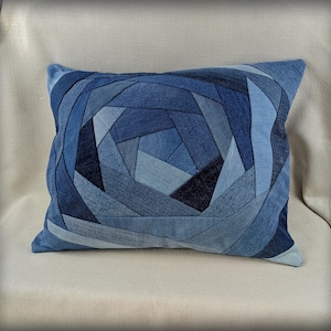 Puede incluir: Cojín cuadrado de mezclilla con un diseño geométrico en tonos azules. El cojín está hecho de mezclilla reciclada, con un patrón de patchwork que crea una superficie texturizada. El diseño presenta una espiral central.