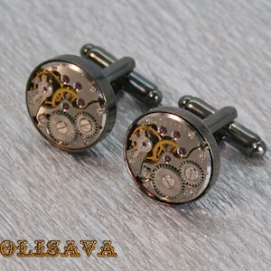 May include: A pair of cufflinks with a black metal finish. Each cufflink features a round, clear glass face with a visible watch movement and small, pink gemstones.