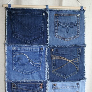 Op de afbeelding: Een wandhanger gemaakt van hergebruikt denim met zes zakken. De zakken zijn allemaal van verschillende grootte en hebben verschillende stikdetails. De denim is donkerblauw en heeft een verweerde look.