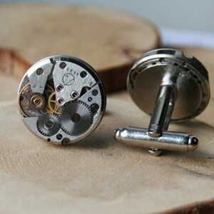 Peut inclure: Une paire de boutons de manchette en argent avec un design circulaire. Chaque bouton de manchette pr&eacute;sente un m&eacute;canisme d'horlogerie d&eacute;taill&eacute; et expos&eacute; avec des engrenages et un fond argent&eacute;.