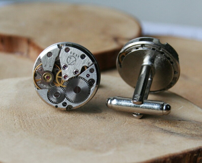 Peut inclure: Une paire de boutons de manchette en argent avec un design circulaire. Chaque bouton de manchette pr&eacute;sente un m&eacute;canisme d'horlogerie d&eacute;taill&eacute; et expos&eacute; avec des engrenages et un fond argent&eacute;.