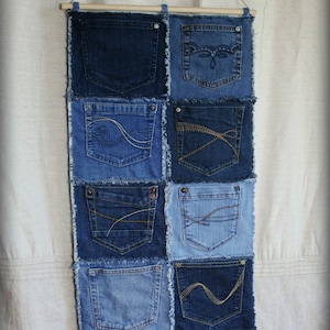 Op de afbeelding: Een wandhanger gemaakt van denim stof met meerdere zakken. De zakken zijn aan elkaar genaaid om een patchwork ontwerp te cre&euml;ren. De stof is lichtblauw van kleur met rafelige randen.