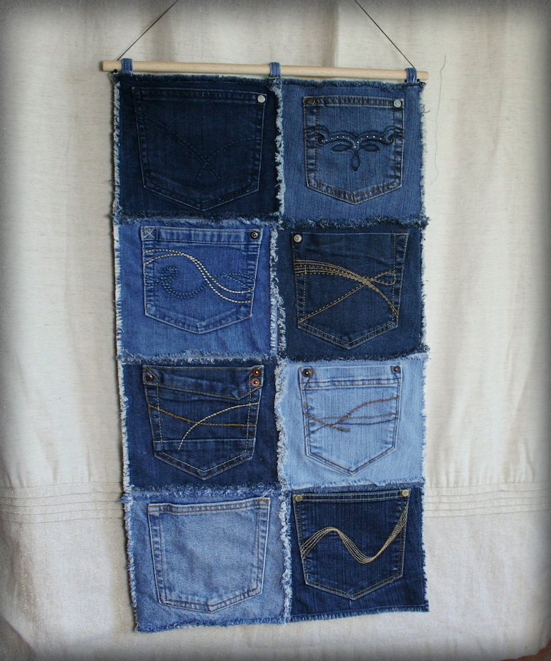 Op de afbeelding: Een wandhanger gemaakt van denim stof met meerdere zakken. De zakken zijn aan elkaar genaaid om een patchwork ontwerp te cre&euml;ren. De stof is lichtblauw van kleur met rafelige randen.